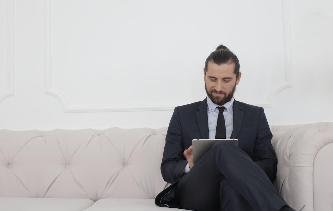 our-story-01 A businessman in a suit sitting on a sofa using a tablet, smiling.